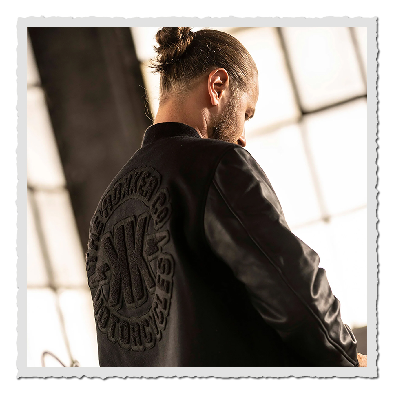 Person in schwarzer Jacke von hinten/seitlich, großes Rückenlogo, Innenraum mit großen Fenstern im Hintergrund.